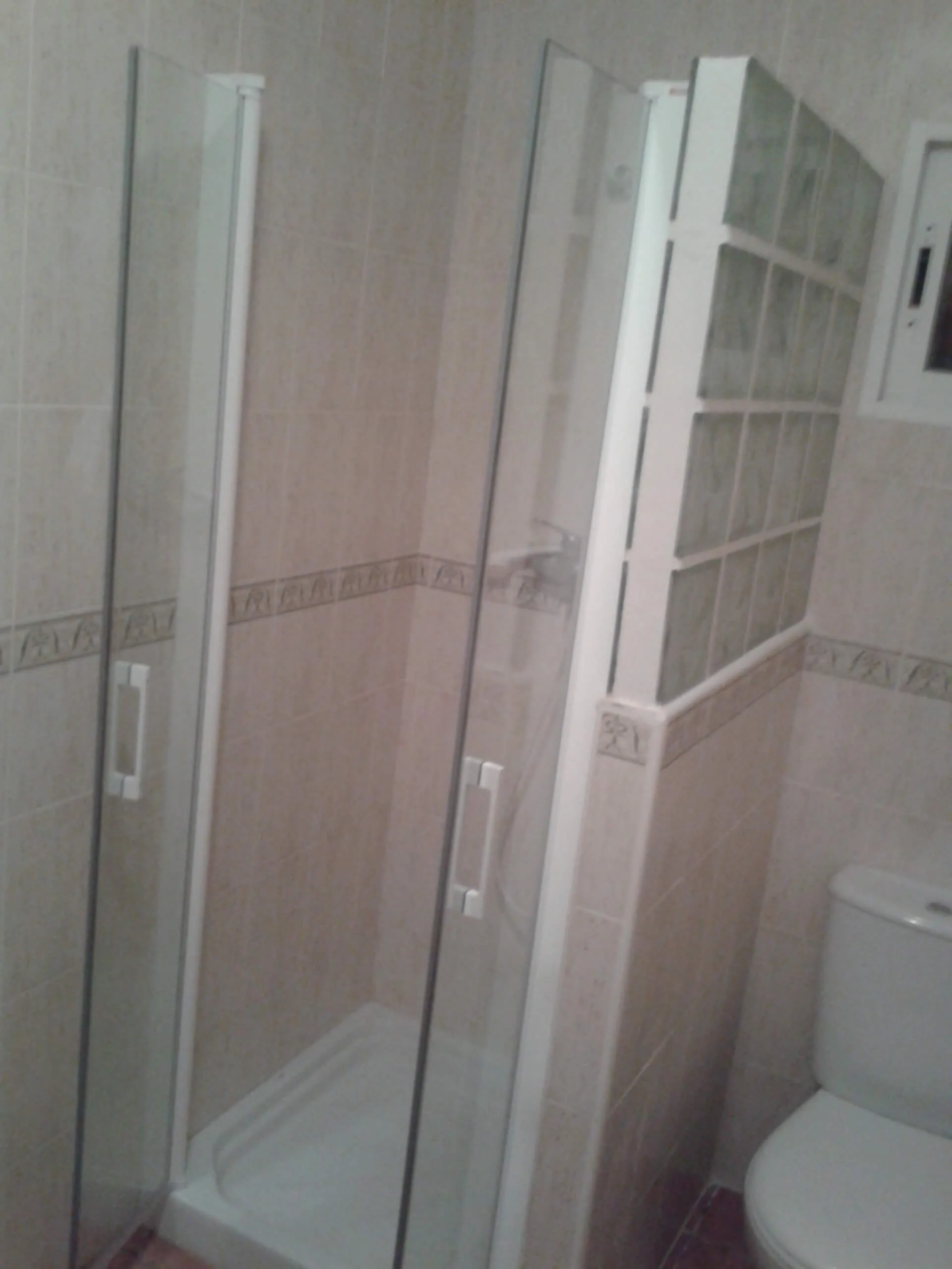 Mampara de ducha abatible en cristal templado transparente con tiradores blancos, instalada en baño pequeño con azulejo beige y separación de pavés, fabricada por Carpintería Metálica Toni en San Pedro, Albacete.