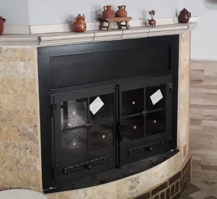 Puerta metálica de chimenea fabricada a medida por Carpintería Metálica Toni, en San Pedro (Albacete). Con doble hoja abatible, cristal resistente al calor y diseño con detalles en forja, mejora la eficiencia térmica y la seguridad del hogar, manteniendo un estilo rústico y elegante.