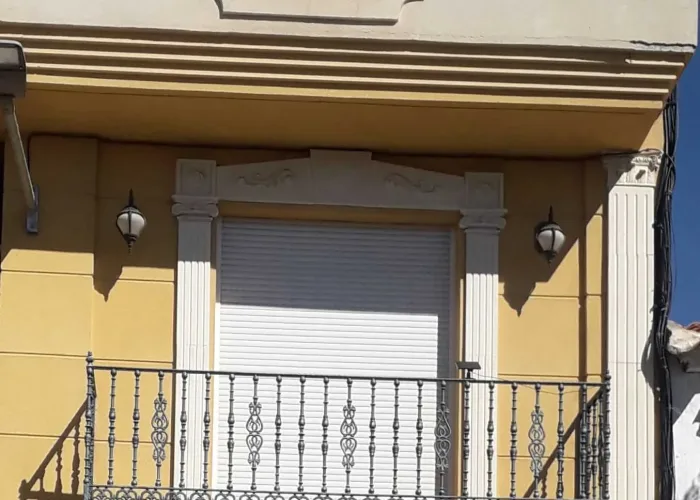 Barandilla de forja artística en balcón clásico con persiana enrollable y molduras ornamentales, instalada por Carpintería Metálica Toni en San Pedro, Albacete.
