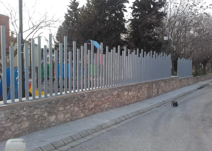 Cerramiento metálico para parque infantil fabricado por Carpintería Metálica Toni en San Pedro, Albacete, con diseño moderno en varilla redonda de diferentes alturas sobre muro de piedra.