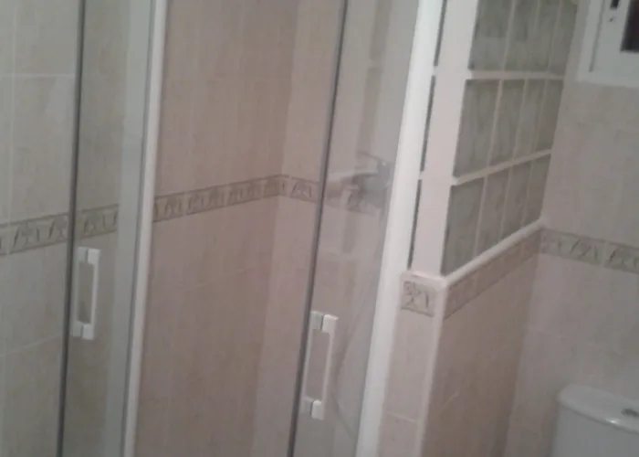 Mampara de ducha abatible en cristal templado transparente con tiradores blancos, instalada en baño pequeño con azulejo beige y separación de pavés, fabricada por Carpintería Metálica Toni en San Pedro, Albacete.