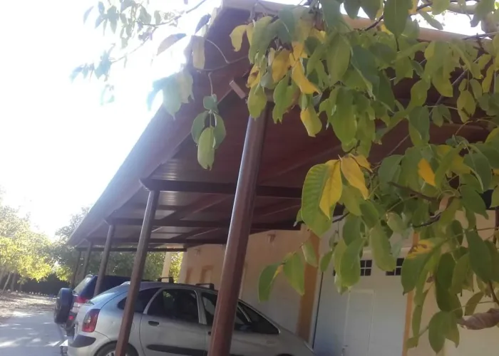 Estructura metálica de cochera con cubierta de panel sándwich imitación madera instalada por Carpintería Metálica Toni en San Pedro, Albacete. Protege varios vehículos en zona exterior junto a vivienda, rodeada de árboles.