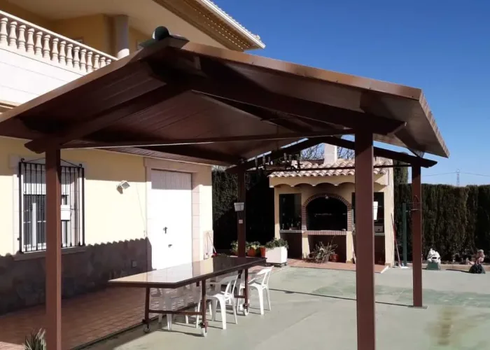 Porche metálico con cubierta de panel sándwich acabado madera, instalado en vivienda unifamiliar. Proyecto realizado por Carpintería Metálica Toni en San Pedro, Albacete, ideal para zonas de comedor exterior o sombra en jardín.