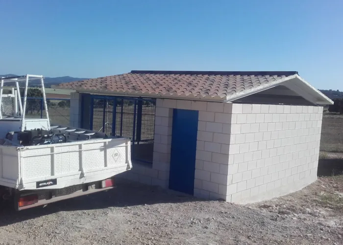 Construcción rural con cerramiento metálico y tejado de panel sándwich imitación teja, realizada por Carpintería Metálica Toni en San Pedro, Albacete. Incluye estructura de bloques de hormigón, puerta metálica azul y cerramiento perimetral con malla electrosoldada.