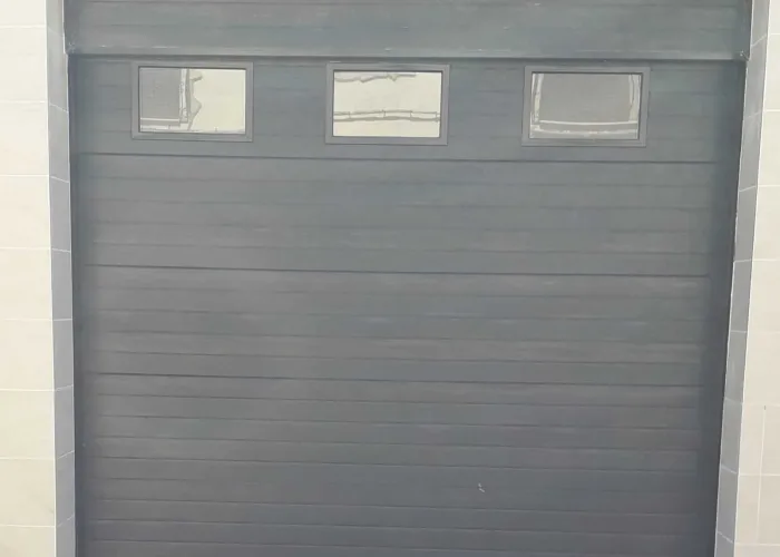 Puerta seccional automática en panel sándwich gris oscuro con ventanas rectangulares superiores, fabricada por Carpintería Metálica Toni en San Pedro, Albacete