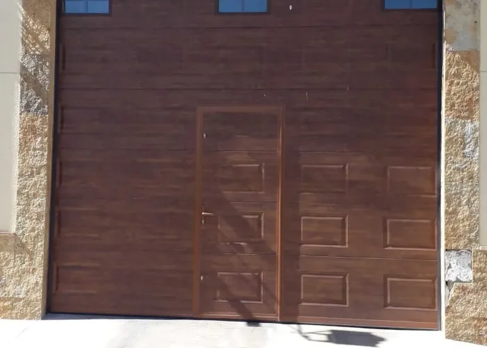 Puerta seccional automatizada de panel sándwich con acabado imitación madera y puerta peatonal integrada, fabricada por Carpintería Metálica Toni en San Pedro, Albacete