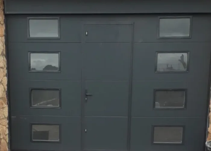 Puerta seccional de panel sándwich en color gris con ventanas acristaladas y puerta peatonal integrada, fabricada por Carpintería Metálica Toni en San Pedro, Albacete