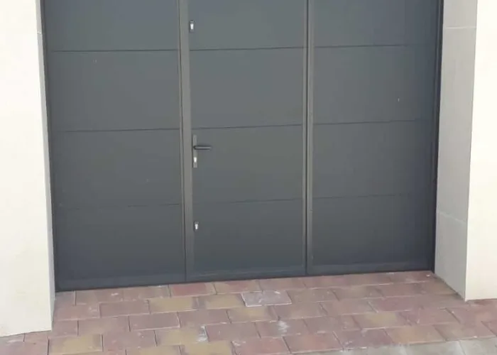 Puerta seccional de garaje en panel sándwich gris con ventanas superiores y puerta peatonal central, fabricada por Carpintería Metálica Toni en San Pedro, Albacete