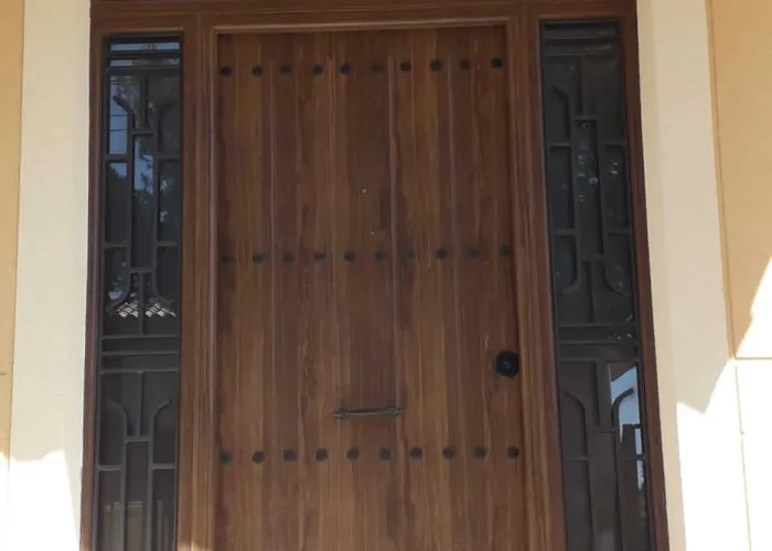 Puerta de entrada en aluminio acabado madera con clavos decorativos y rejas laterales y superiores forjadas, fabricada por Carpintería Metálica Toni en Albacete