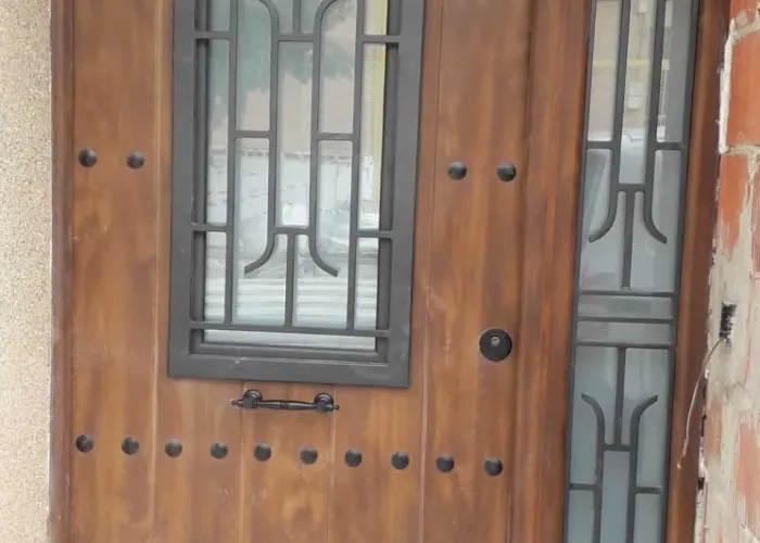 Puerta de entrada de aluminio imitación madera con clavos decorativos, ventana central y rejas forjadas, fabricada por Carpintería Metálica Toni en San Pedro, Albacete