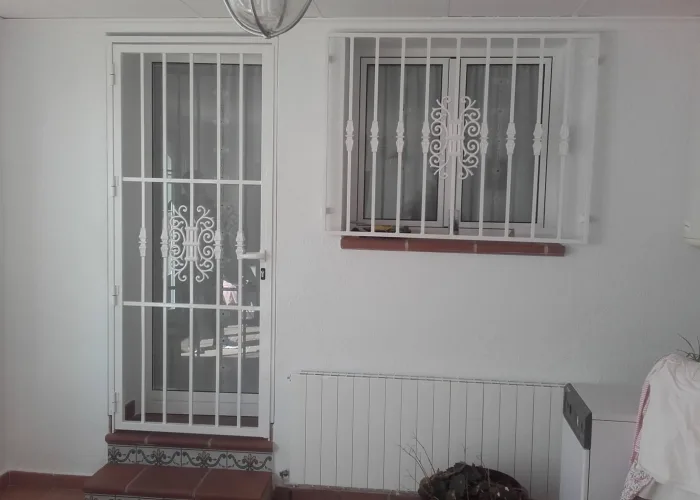 Rejas de seguridad blancas para puerta y ventana, fabricadas en hierro por Carpintería Metálica Toni en San Pedro (Albacete). Diseño con barrotes verticales y detalle ornamental central, ideales para interiores o patios cerrados que requieren protección sin renunciar a la estética.