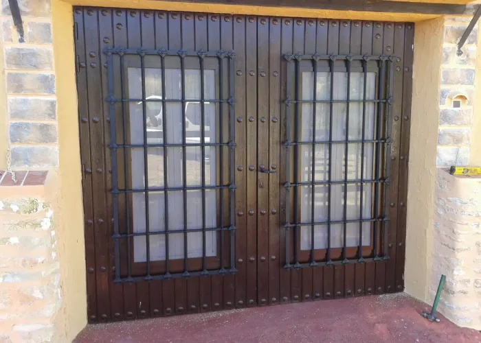 Puerta de doble hoja en madera oscura con rejas metálicas frontales de diseño clásico, fabricada por Carpintería Metálica Toni en San Pedro (Albacete). Una combinación robusta y estética para entradas principales de casas rústicas o viviendas con encanto tradicional.